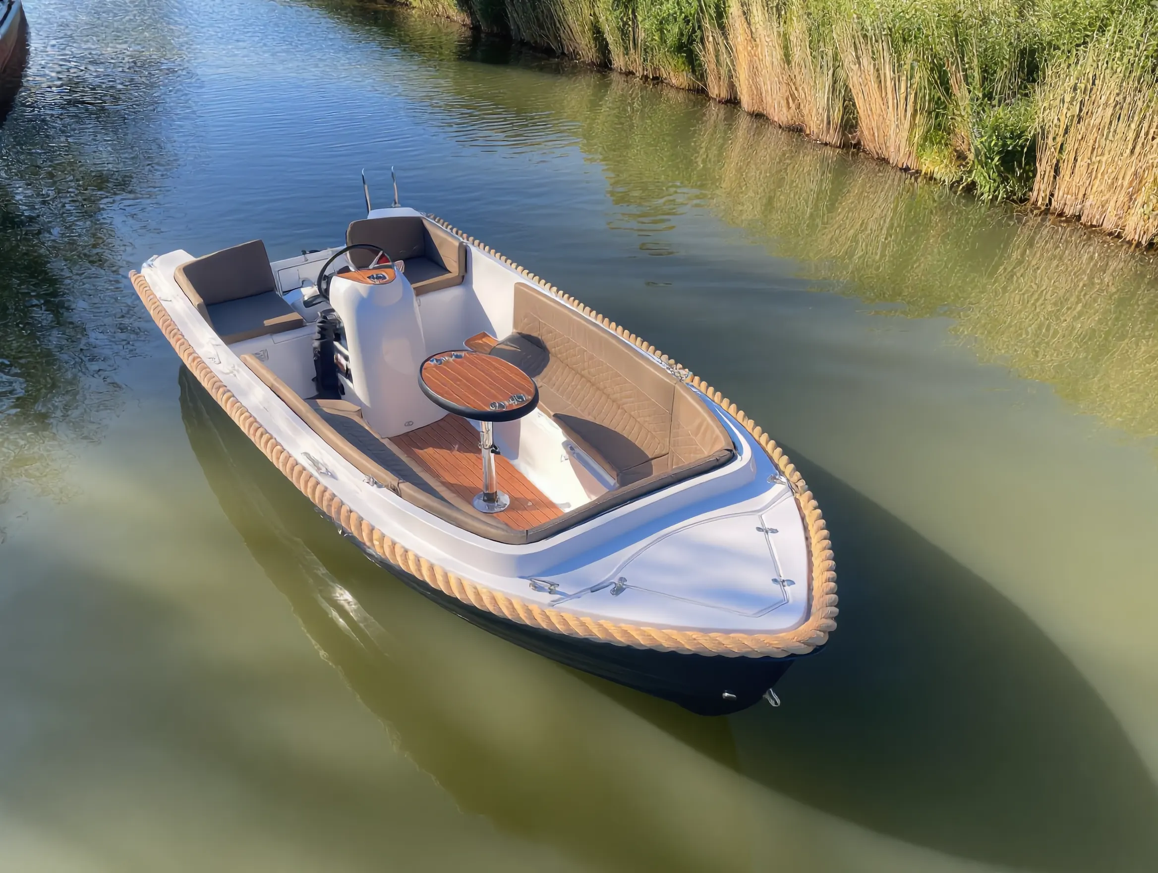 Onze sloep met teakdek, ronde tafel en beige kussenbanken, stil liggend op rustig water langs een rietkraag